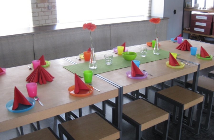 Ein langer Tisch ist mit bunten Plastiktellern und -bechern, roten Servietten und zwei kleinen Blumenvasen geschmückt, unter dem Tisch stehen Hocker.
