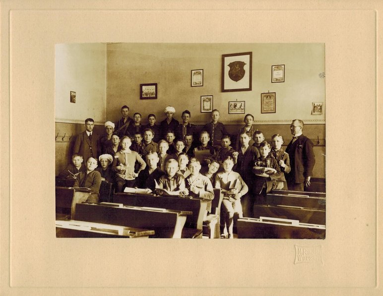 Historisches Foto in Sepia-Tönen, auf dem eine Gruppe von Jungen in einem Klassenzimmer mit Holzbänken steht