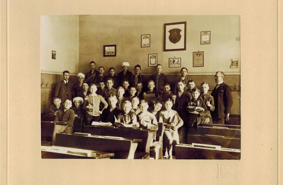Historisches Foto in Sepia-Tönen, auf dem eine Gruppe von Jungen in einem Klassenzimmer mit Holzbänken steht