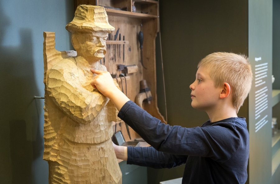 In einem Ausstellungsraum berührt ein Junge die aus Holz geschnitzte, nahezu lebensgroße Figur eines Jägers.