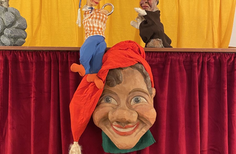 Vor dem geschlossen Vorhang eines Puppentheaters blickt die Figur des Kaspers direkt in die Kamera. Hinter dem Kasper sind zwei weitere Handpuppen zu sehen.