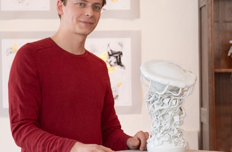Ein junger Mann steht vor einem Kunstwerk aus weißem Porzellan und lächelt in die Kamera.