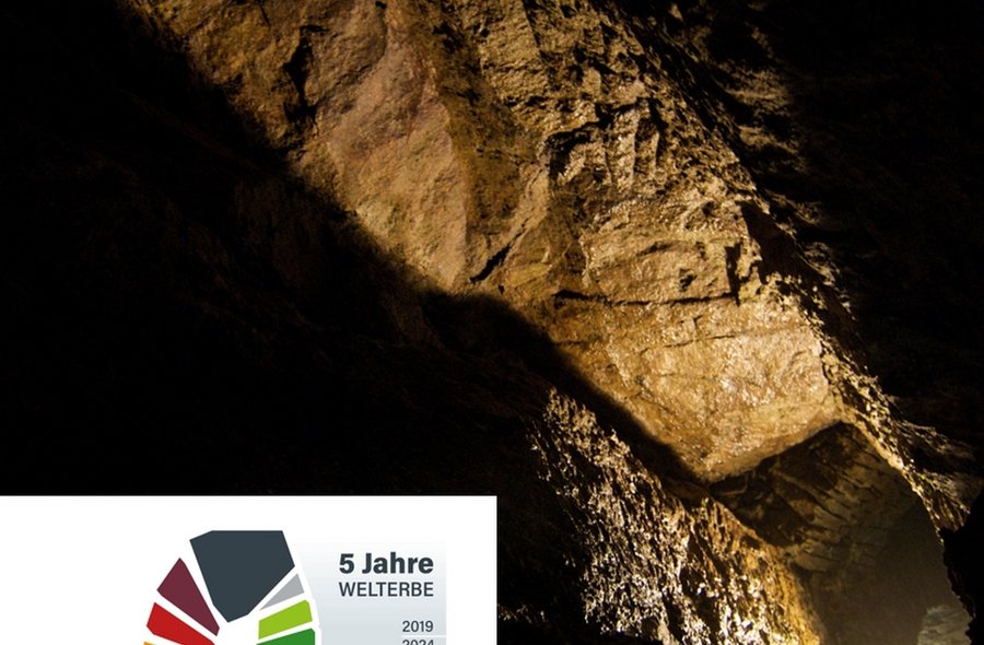 Das Foto gibt den Blick frei in einen beleuchteten Bergwerksstollen. Unten links die Aufschrift "5 Jahre Welterbe Montanregion Erzgebirge/Krušnohoří, 2019 bis 2024".
