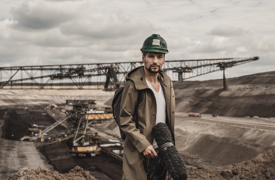 Ein Mann mit braunem Regenmantel und grünem Bauarbeiter-Helm steht, ein großes Mikrofon für Filmproduktionen haltend, in einer Tagebaulandschaft mit Abbaubagger im Hintergrund.