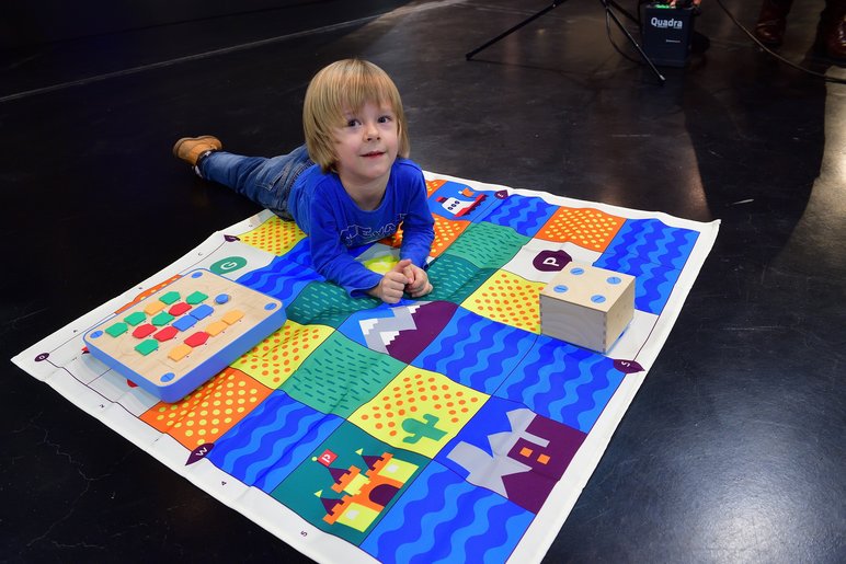 Ein kleines Kind liegt auf einem bunten Spielteppich und blickt verschmitzt in die Kamera.