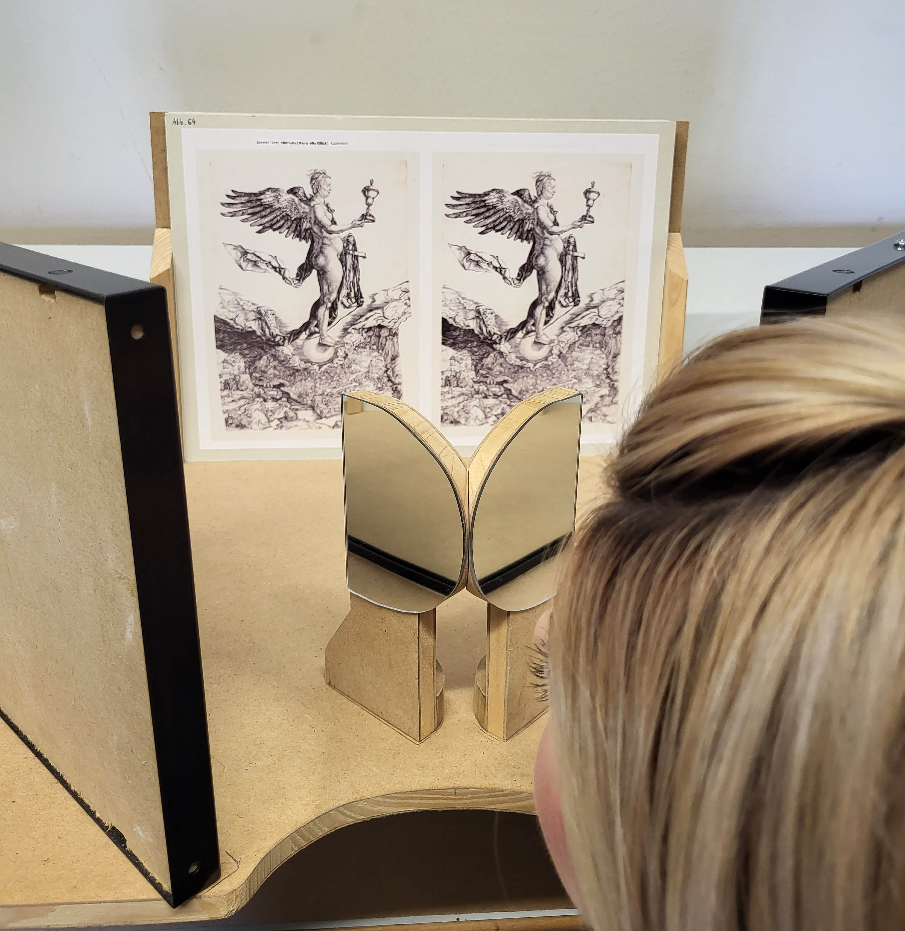 Eine von hinten zu sehende Person blickt in Richtung eines auf einem Tisch angeordneten stereoskopischen Doppelbildes.