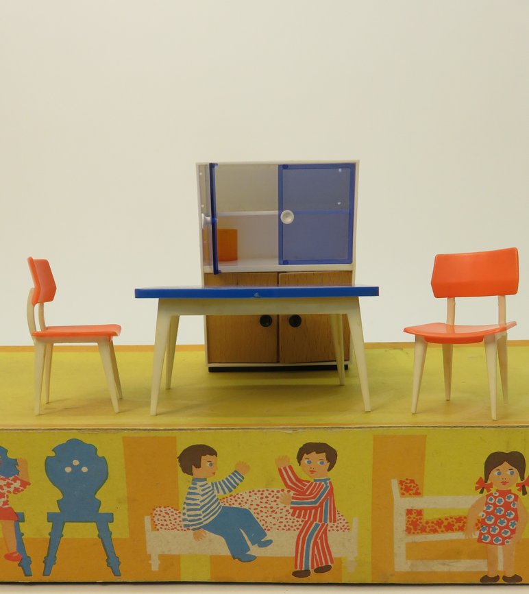 Zwei Miniaturstühle und ein Miniaturtisch stehen vor einem Miniaturschrank. Das gelbe Podest, auf dem die Puppenmöbel stehen, ist mit Zeichnungen von Puppenkindern in einer Puppenstube verziert.