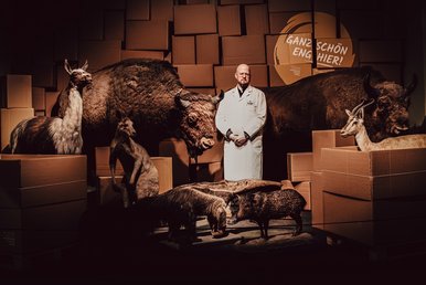 „Dr.“ W. Astersen im Naturkundemuseum Leipzig Ein Mann mit Glatze, Bart und weißem Kittel steht inmitten einer Inszenierung größerer und kleinerer ausgestopfter Wildtiere.