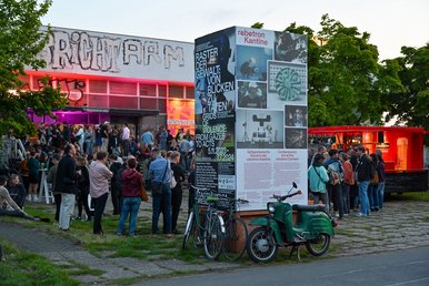 robotron-Kantine 2024 Viele Menschen an einem Sommerabend vor einem mit Graffiti versehenen Bau der Ostmoderne, davor ein mit Plakaten beklebter Pfeiler, an dem ein Fahrrad und ein Moped lehnen