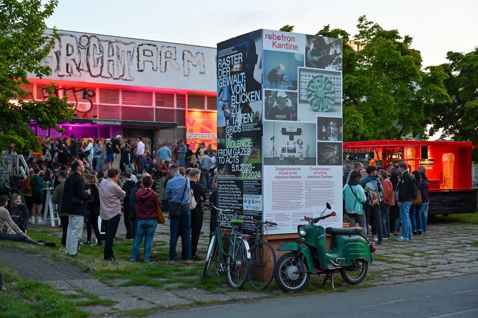Viele Menschen an einem Sommerabend vor einem mit Graffiti versehenen Bau der Ostmoderne, davor ein mit Plakaten beklebter Pfeiler, an dem ein Fahrrad und ein Moped lehnen