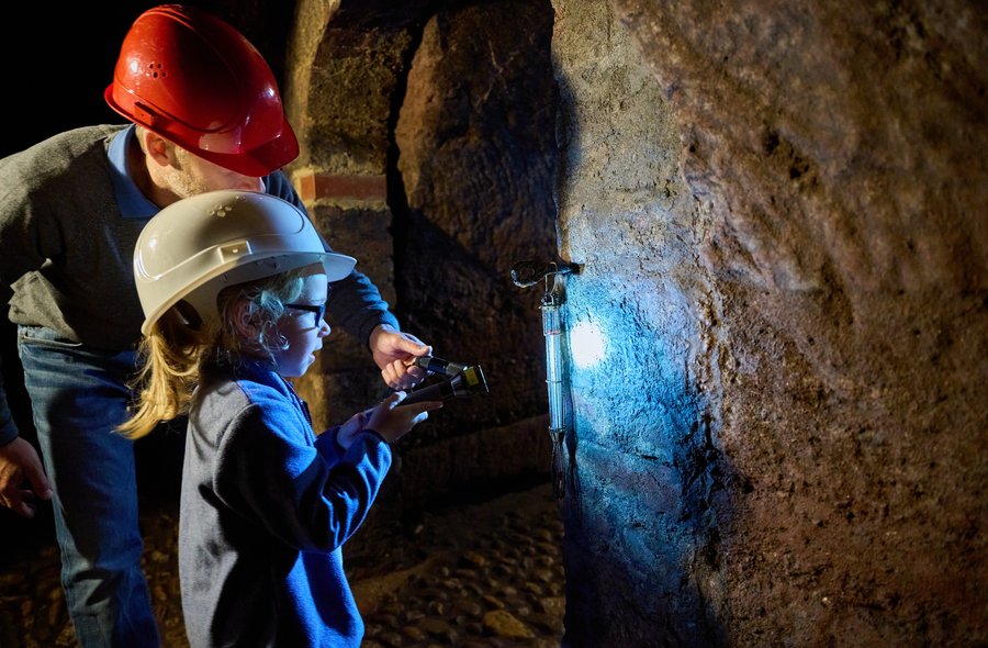 Taschenlampenführung in unterirdischen Gängen Ein Erwachsener und ein Kind, beide mit Helm, stehen in einem dunklen historischen Kellergang und leuchten mit einer Taschenlampe ein Objekt an einer Wand an.