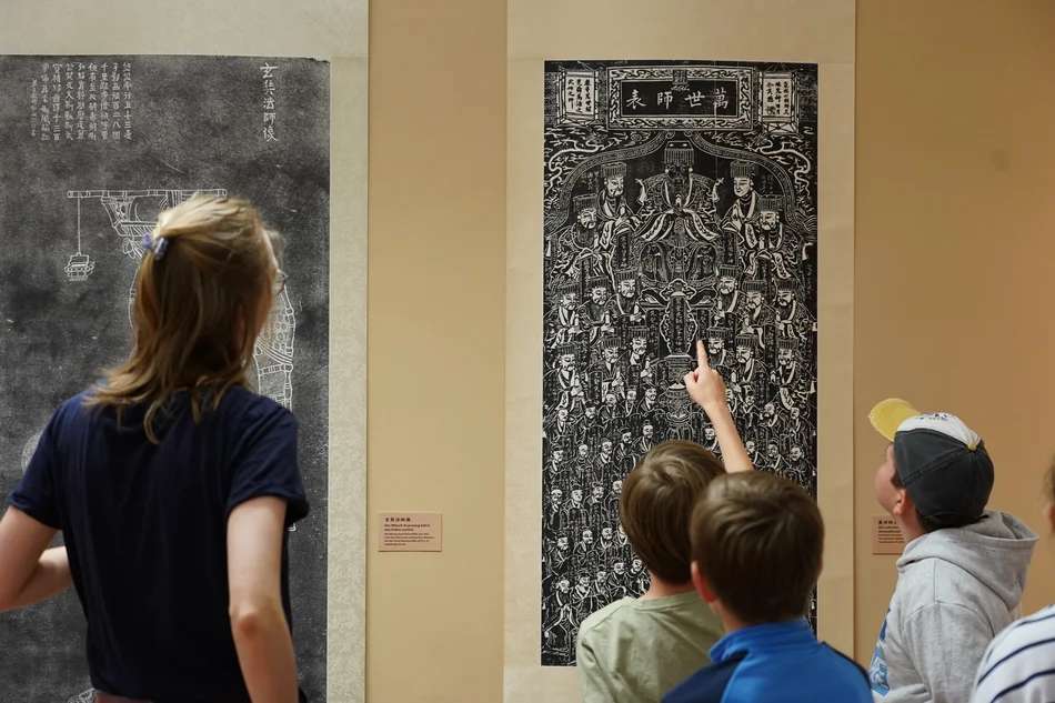 Drei Kinder und eine Erwachsene betrachten zwei großformatige chinesische Druckgrafiken.