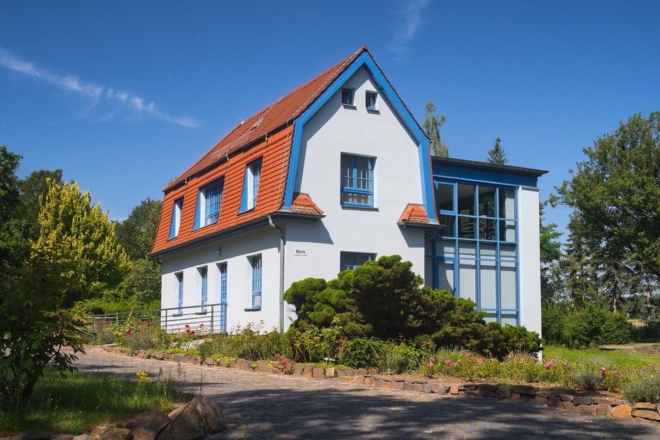 Weißes historisches Haus mit modern wirkendem Anbau, umgeben von Bäumen, Blumenrabatten und Weg