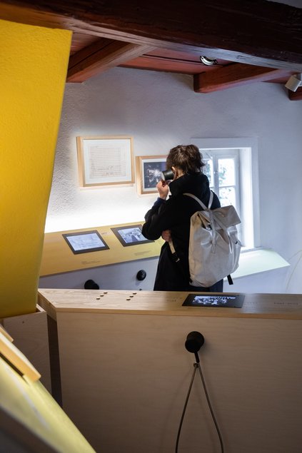 In einem Ausstellungsraum stehen im spitzen Winkel zueinander angeordnet zwei Medienstationen. Dazwischen steht eine Frau, die sich den Hörer einer Hörstation ans Ohr hält.