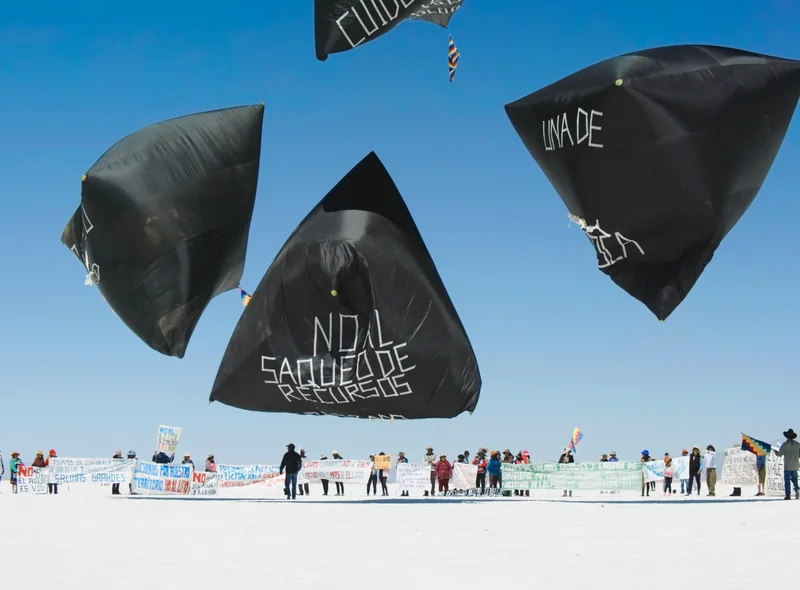 Auf weißem, flachen Boden mehrere Menschen, darüber schweben riesige, mit Schriftzügen versehene, pyramidenförmige Ballons.