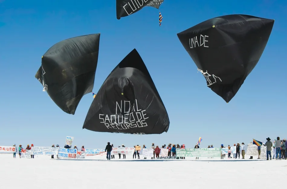 Tomás Saraceno, „Fly with Pacha, Into the Aerocene“, 2017–2023, 1-Kanal-Video, 76:25 min Auf weißem, flachen Boden mehrere Menschen, darüber schweben riesige, mit Schriftzügen versehene, pyramidenförmige Ballons.