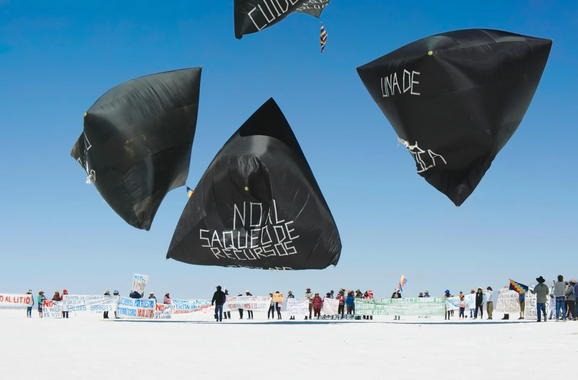 Tomás Saraceno, „Fly with Pacha, Into the Aerocene“, 2017–2023, 1-Kanal-Video, 76:25 min Auf weißem, flachen Boden mehrere Menschen, darüber schweben riesige, mit Schriftzügen versehene, pyramidenförmige Ballons.