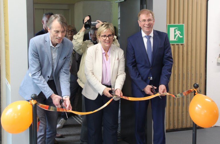 Eine Frau und zwei zu ihrer Linken und zu ihrer Rechten stehende Männer durchtrennen mit Scheren ein orangefarbenes Band, an dem zwei orangefarbene Luftballons befestigt sind.