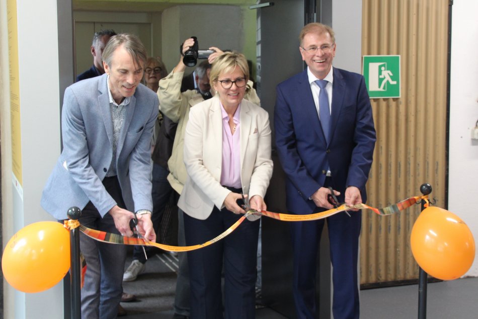 Eine Frau und zwei zu ihrer Linken und zu ihrer Rechten stehende Männer durchtrennen mit Scheren ein orangefarbenes Band, an dem zwei orangefarbene Luftballons befestigt sind.