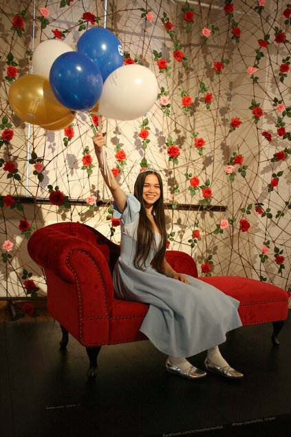 Eine in ein graues Dornröschen-Kleid gekleidete junge Frau sitzt auf einer roten Récamière und hält Luftballons hoch. Die Wand im Hintergrund schmückt eine Rosenhecke.
