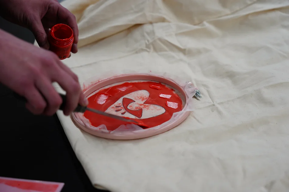 Durchlässiges Gewebe ist in einen runden Stickrahmen eingespannt, darauf ist ein verspieltes Motiv erkennbar. Eine Person bestreicht das Gewebe mit roter Farbe.