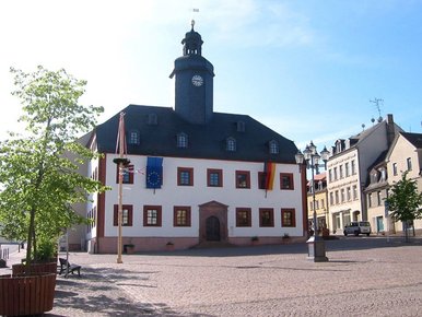 Sommerliche Außenansichtes eines dreistöckigen weißen Gebäudes mit schwarzem Schieferdach und einem zentral angeordneten schwarzen Türmchen, das am Rande eines großen, Kopfstein-gepflasterten Platzes steht
