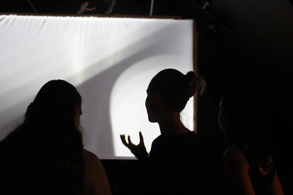 Drei Personen, von hinten gesehen, stehen in einem dunklen Raum vor einer großen, weißen Leinwand.