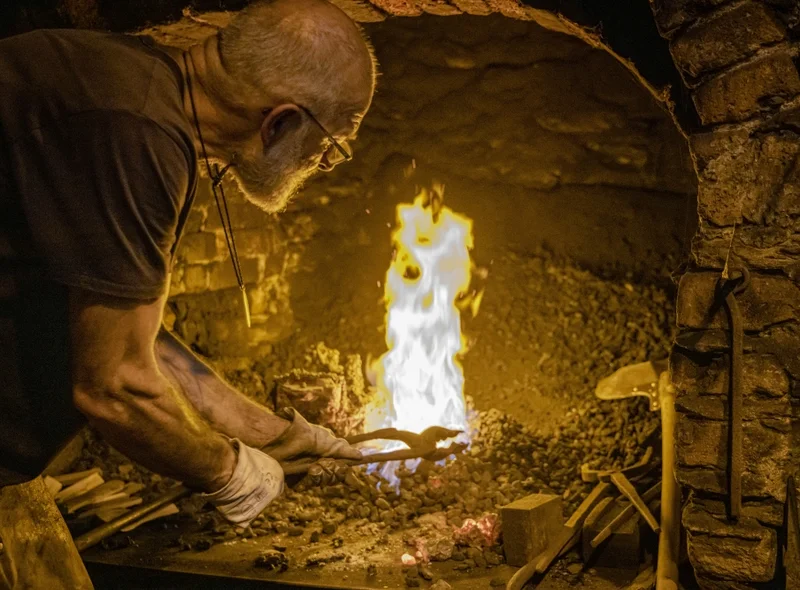 Ein Schmied führt eine eiserne Zange in ein loderndes Feuer ein.