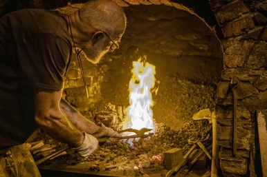 Schauschmieden Ein Schmied führt eine eiserne Zange in ein loderndes Feuer ein.