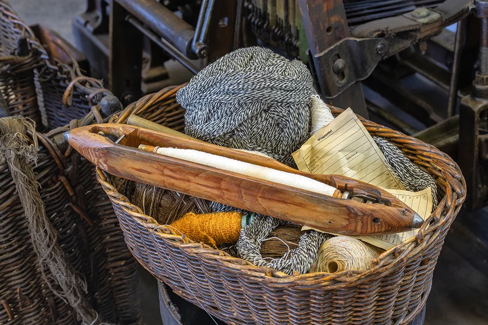 Weberei: Korb mit Weberschiffchen In einem Bastkörbchen liegen Garne und ein hölzernes Schiffchen-förmiges Objekt, in dem eine Garnspindel steckt.