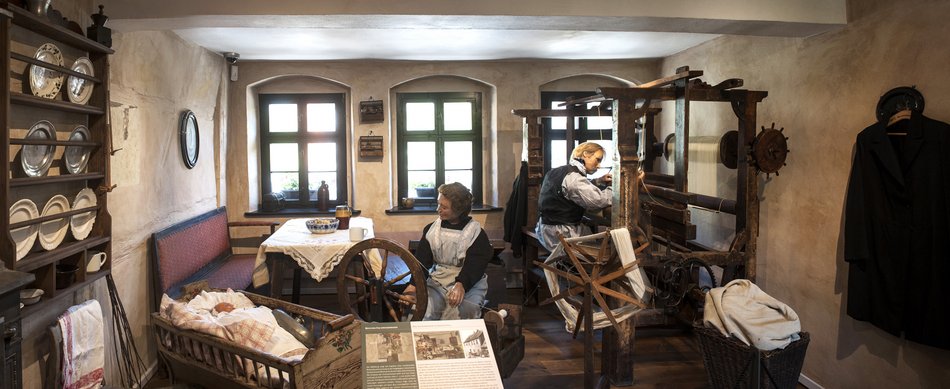 Das Foto zeigt eine Inszenierung der von Armut bestimmten Wohnsituation der Familie May: die Weberstube im historischen Geburtshaus Karl Mays. In dem beengten Raum wird gewohnt wie auch gearbeitet: eine männliche Figur sitzt an einem Webstuhl, eine weibliche Figur an einem Spinnrad. Neben ihr liegt die Figur eines Babys in einer Wiege, hinter ihr befinden sich ein Esstisch und eine Sitzbank. im Regal an der linken Wand befinden sich Teller, darunter hängt ein Geschirrhandtuch.