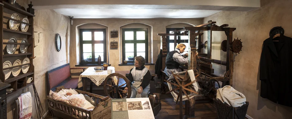 Weberstube im Karl-May-Haus Das Foto zeigt eine Inszenierung der von Armut bestimmten Wohnsituation der Familie May: die Weberstube im historischen Geburtshaus Karl Mays. In dem beengten Raum wird gewohnt wie auch gearbeitet: eine männliche Figur sitzt an einem Webstuhl, eine weibliche Figur an einem Spinnrad. Neben ihr liegt die Figur eines Babys in einer Wiege, hinter ihr befinden sich ein Esstisch und eine Sitzbank. im Regal an der linken Wand befinden sich Teller, darunter hängt ein Geschirrhandtuch.