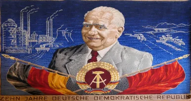 In Teppich geknüpftes Porträt des Präsidenten der DDR vor Industriebauten, darunter die Flagge der DDR und eine rote Arbeiterfahne