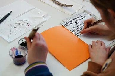 Nahaufnahme zweier Kinderhände, die mit Federkiel und Tinte in ein Schreibheft schreiben.