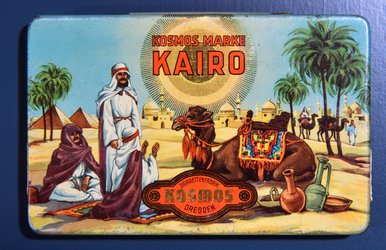 Das Foto zeigt eine Zigarettendose der Marke „Kairo“, auf der orientalisch anmutende Personen sowie Kamele und Palmen vor den Pyramiden von Gizeh abgebildet sind.