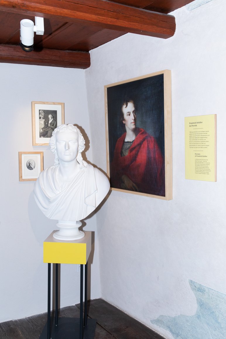 In der Ecke eines Ausstellungsraumes steht eine weiße Büste Friedrich Schillers, an den Wänden dahinter hängen eine Porträtmalerei und zwei kleinere Grafiken. Den oberen Abschluss des Raumes bildet eine hölzerne Balkendecke.