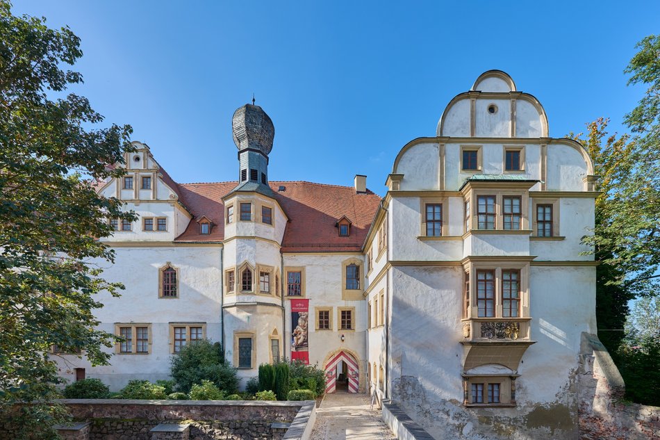 Sommerliche Ansicht einer von Bäumen umgebenen weiß getünchten Schlossfassade unter blauem Himmel