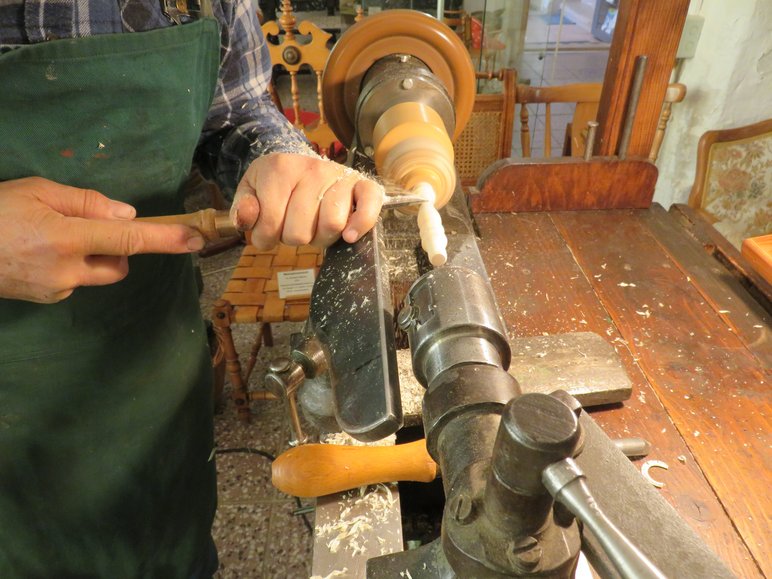 Ein Holzobjekt ist in eine Drechselmaschine eingespannt und dreht sich schnell, dabei drückt ein Handwerker mit einem Werkzeug gegen das Objekt, um es zu formen.