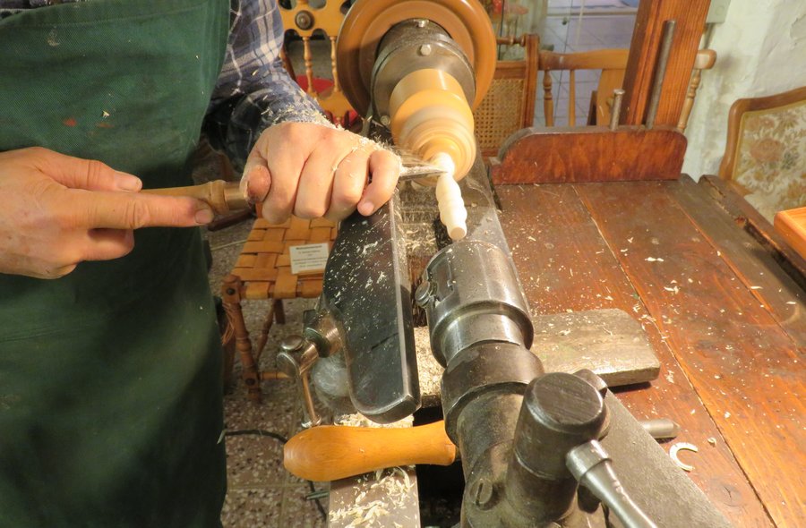 Ein Holzobjekt ist in eine Drechselmaschine eingespannt und dreht sich schnell, dabei drückt ein Handwerker mit einem Werkzeug gegen das Objekt, um es zu formen.