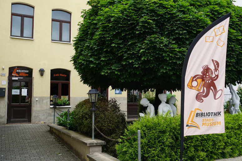 Die Außenansicht zeigt den Eingang zum Gebäude von Bibliothek und Stadtmuseum Pulsnitz. Auf dem Fenster neben der Eingangstür steht der Schriftzug "Café Marie". Neben dem Fußweg zur Eingangstür befindet sich eine Grünfläche mit Büschen, einem Baum und mehreren weißen Skulpturen. Im Vordergrund weht auf einem Aufsteller die Fahne von Bibliothek und Stadtmuseum, von der Cartoon-artig ein Lebkuchenmännchen grüßt.