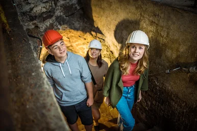 Zwei junge Frauen mit weißen Helmen und ein junger Mann mit rotem Helm steigen einen tunnelartigen, in Fels gehauenen Gang hinauf.