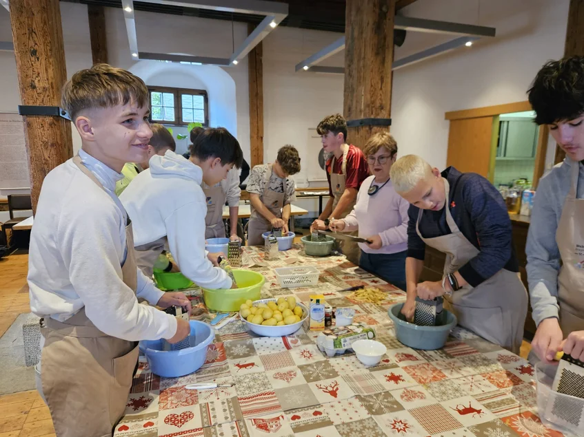 Gemeinsames Kartoffelreiben an einem langen Tisch; Teilnehmende mit Schürzen arbeiten konzentriert.