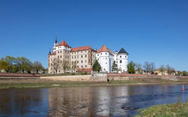 Das Foto zeigt eine sommerliche Panorama-Ansicht von Schloss Hartenfels in Torgau, fotografiert vom gegenüberliegenden Ufer der Elbe aus.