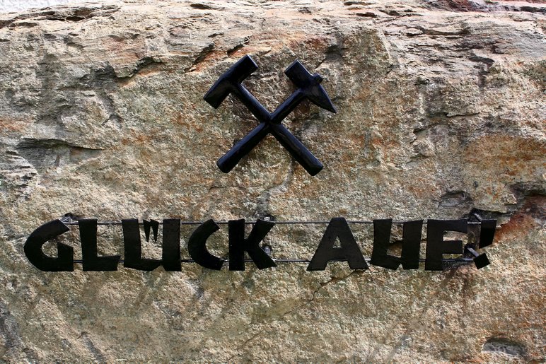 Schwarzer Schriftzug "Glück Auf" und Eisen und Schlägel Symbolik auf hellgrauem Stein