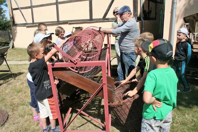 Zwei Erwachsene stehen mit einer Grundschulgruppe im Außenbereich des Museums. Ein Erwachsener erklärt das historische Kartoffelwäsche-Gerät.