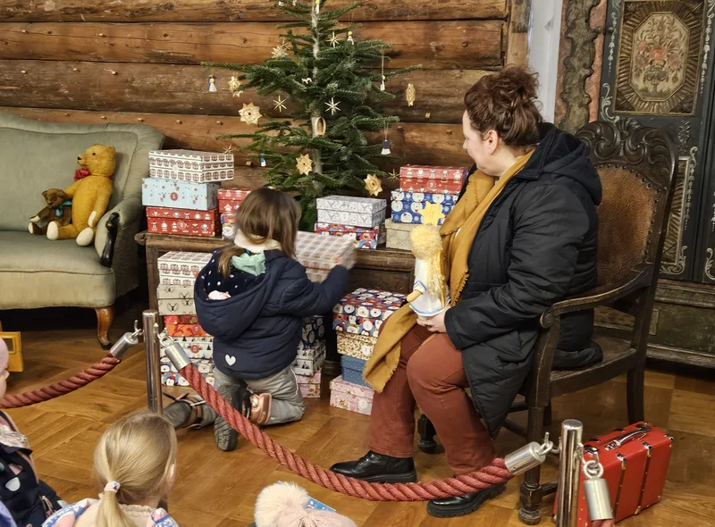 Ein Kind kniet vor einem geschmücktem Tannenbaum und Weihnachtsgeschenken, ein Frau daneben schaut zu.