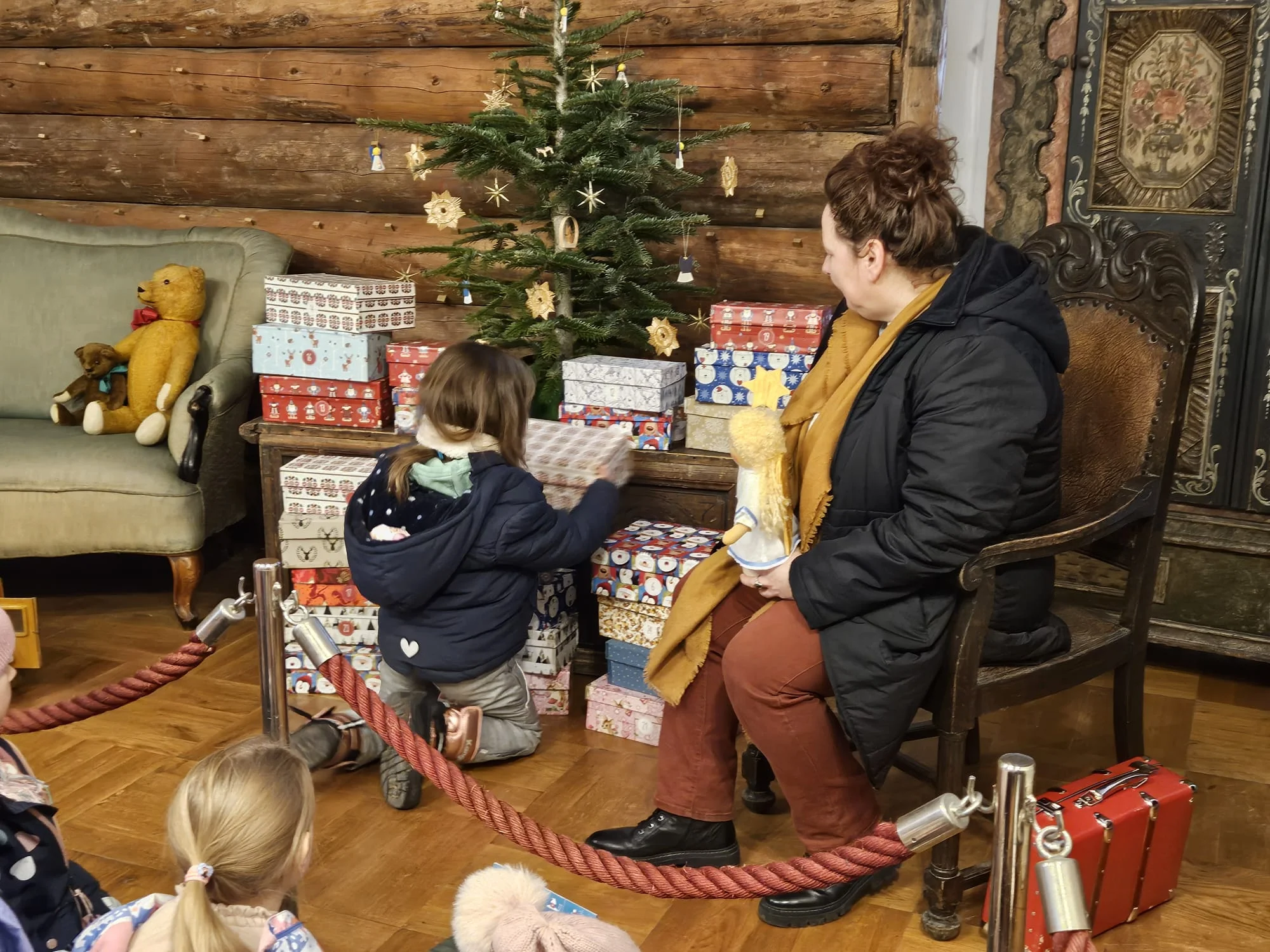 Ein Kind kniet vor einem geschmücktem Tannenbaum und Weihnachtsgeschenken, ein Frau daneben schaut zu.