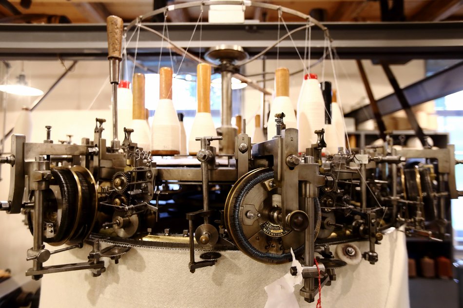 Das Foto zeigt eine Detailaufnahme eines historischen Rundkulierwirkstuhls mit seiner komplexen Mechanik und mehreren weißen Garnspulen.