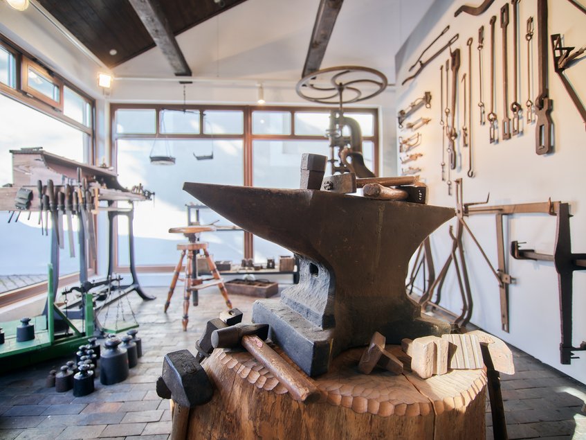 Foto aus dem Inneren der im Oschatzer Waagenmuseum inszenierten historischen Waagenbauwerkstatt, mit einem Amboss im Vordergrund, einer Werkbank im Hintergrund und einer Vielzahl unterschiedlichster Werkzeuge und Gewichte im Raum und an den Wänden