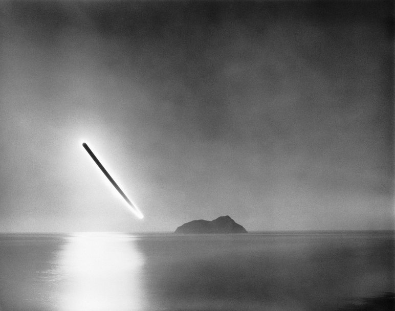 Langzeitbelichtete Schwarzweißfotografie eines Sonnenaufgangs über einer Wasserfläche mit einer zentral im Bild befindlichen Insel